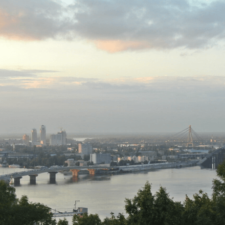 A picture of Kyiv, Ukraine with many buildings, a river, a bridge, many trees, and the cloudy sky