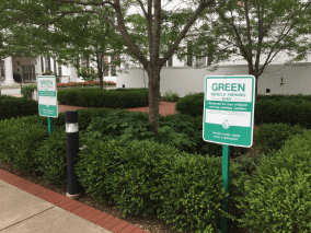 A picture of a small road in a city with two signs, green bushes, trees, and houses