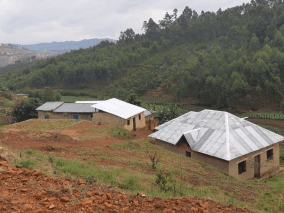 A picture of the mountainous countryside with red lands, two small houses, green trees, and the cloudy sky